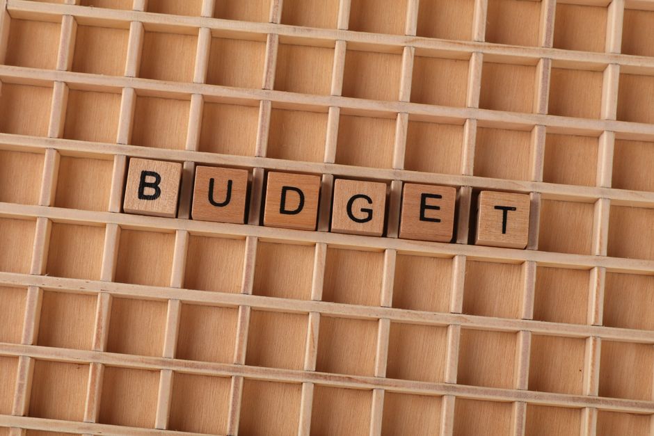 A close-up of a wooden grid tray with multiple empty compartments, placed on a flat surface. In the center of the tray, five wooden letter tiles spell out the word 'BUDGET' in black capital letters. T
