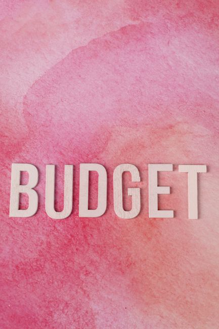 A close-up of a wooden grid tray with multiple empty compartments, placed on a flat surface. In the center of the tray, five wooden letter tiles spell out the word 'BUDGET' in black capital letters. T