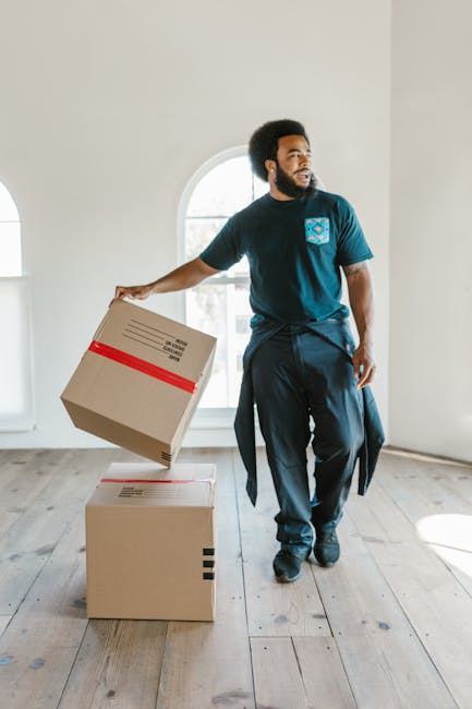 Two professional movers from Removal Companies Barnet are indoors during a home relocation, standing on a light wooden floor in front of large, arched windows allowing natural daylight to fill the roo